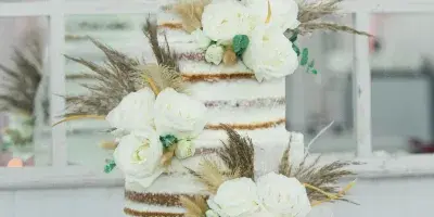 A three-tiered semi-naked wedding cake decorated with white roses, dried grasses, and a gold topper, displayed on a wooden barrel stand.