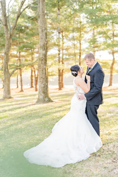 A bride in a white lace gown and a groom in a dark suit standing together in a grassy area with trees in the background.