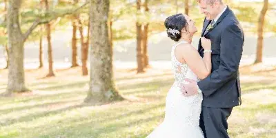 A bride in a white lace gown and a groom in a dark suit standing together in a grassy area with trees in the background.