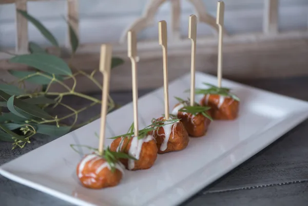 A row of skewered appetizers with sauce and herb garnish on a white rectangular plate.