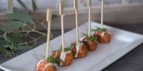 A row of skewered appetizers with sauce and herb garnish on a white rectangular plate.
