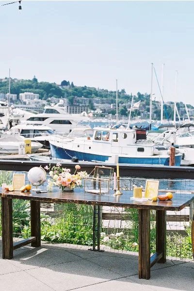 Outdoor wedding welcome table with floral arrangements and gold accents on a waterfront patio, overlooking a marina with boats and a city skyline on a sunny day.
