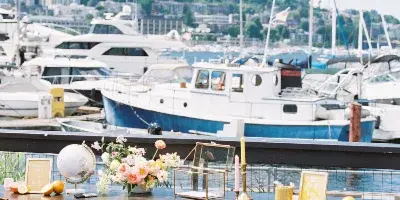 Outdoor wedding welcome table with floral arrangements and gold accents on a waterfront patio, overlooking a marina with boats and a city skyline on a sunny day.