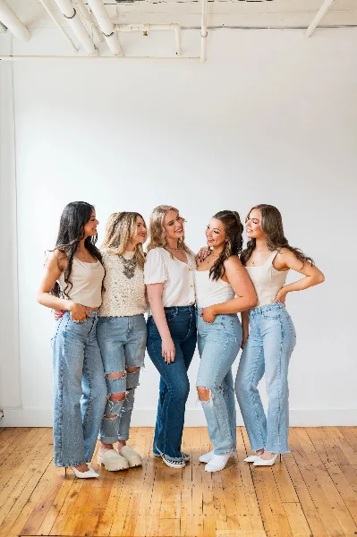 Five smiling women standing together in a bright studio with white walls and wooden floors, wearing casual tops and jeans.
