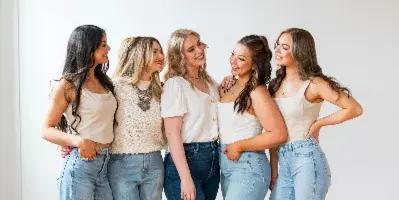 Five smiling women standing together in a bright studio with white walls and wooden floors, wearing casual tops and jeans.