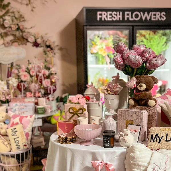 Interior of a floral boutique featuring a 'FRESH FLOWERS' sign, pink rose arrangements, and a display table with gifts and decor.