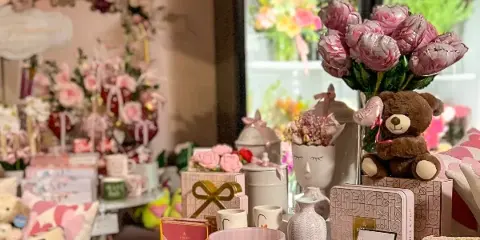 Interior of a floral boutique featuring a 'FRESH FLOWERS' sign, pink rose arrangements, and a display table with gifts and decor.