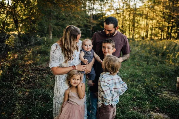 A family of five posing together in a grassy field with a forest background.