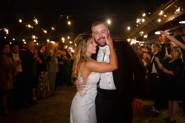 A bride and groom smiling and embracing during a wedding reception with guests holding sparklers in the background.