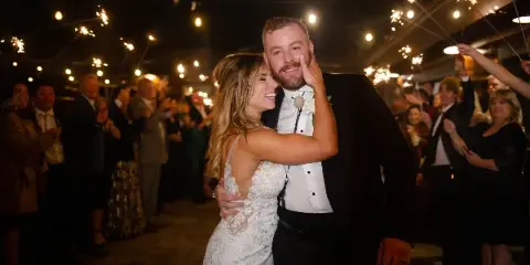 A bride and groom smiling and embracing during a wedding reception with guests holding sparklers in the background.