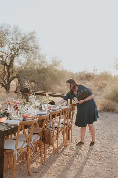 A woman in a dark dress arranging a table setting with floral centerpieces and glassware at an outdoor event venue.
