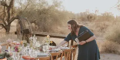 A woman in a dark dress arranging a table setting with floral centerpieces and glassware at an outdoor event venue.