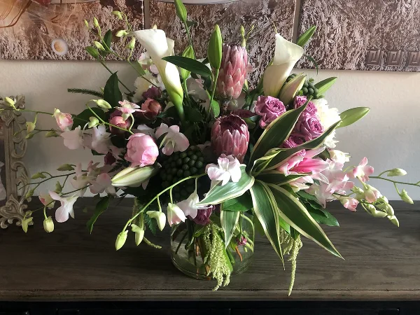 A professional floral arrangement featuring white calla lilies, pink proteas, and orchids in a glass vase on a wooden surface.