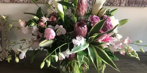 A professional floral arrangement featuring white calla lilies, pink proteas, and orchids in a glass vase on a wooden surface.