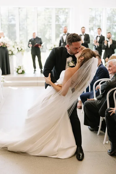 A bride and groom share a kiss during their wedding ceremony in a bright, modern venue with guests seated in the background.