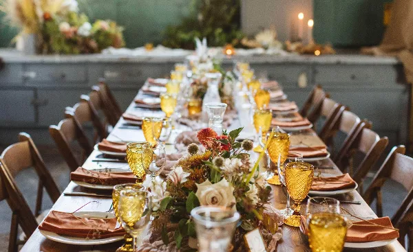 A long event table set with wooden chairs, amber glassware, napkins, and floral centerpieces.