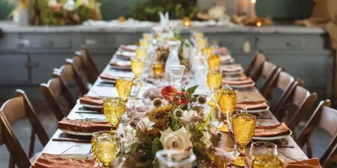 A long event table set with wooden chairs, amber glassware, napkins, and floral centerpieces.