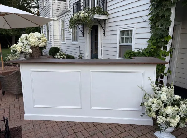 White paneled mobile bar with a dark wood countertop set up on a brick patio with white floral arrangements.