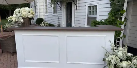 White paneled mobile bar with a dark wood countertop set up on a brick patio with white floral arrangements.