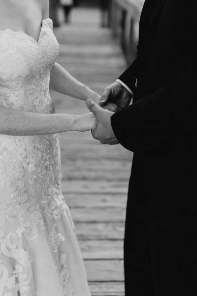 A black-and-white photograph of a bride in a lace gown and a groom in a dark suit holding hands.