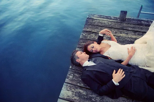 A bride and groom lying on a wooden dock next to a body of water.