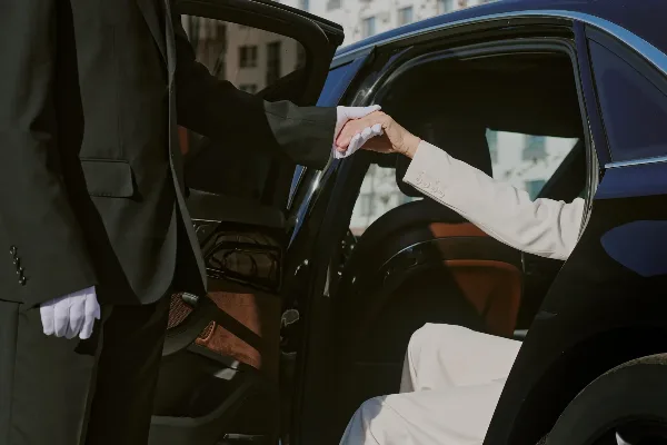 A white-gloved chauffeur assisting a passenger from a black luxury sedan.
