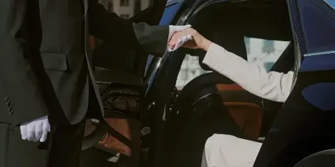 A white-gloved chauffeur assisting a passenger from a black luxury sedan.