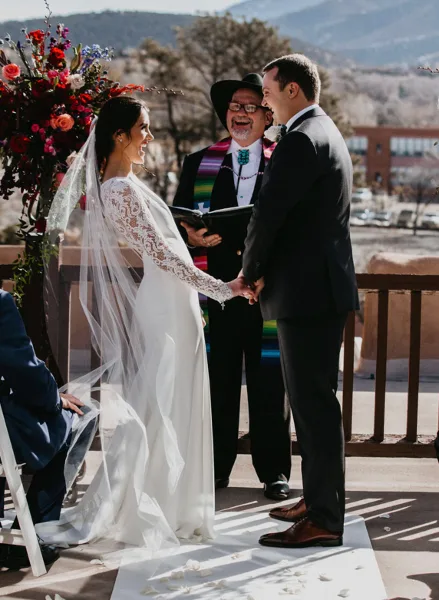 A wedding officiant in a hat and vest officiates a ceremony between a bride and groom outdoors.