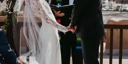 A wedding officiant in a hat and vest officiates a ceremony between a bride and groom outdoors.