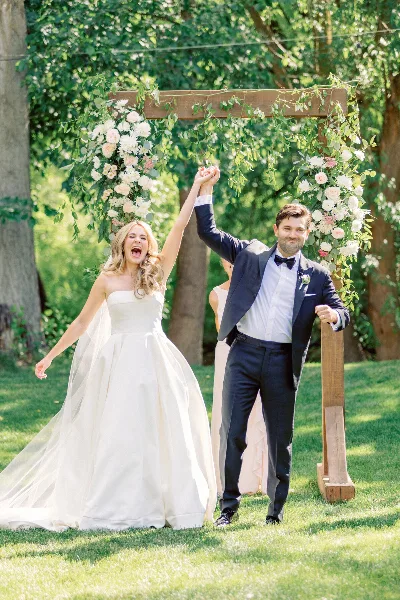 A bride and groom holding hands and smiling outdoors in front of a floral wedding arch.