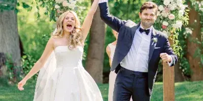A bride and groom holding hands and smiling outdoors in front of a floral wedding arch.