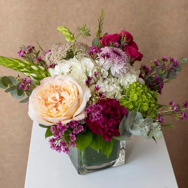 A mixed floral arrangement featuring peach roses, white hydrangeas, and purple accents in a square glass vase.