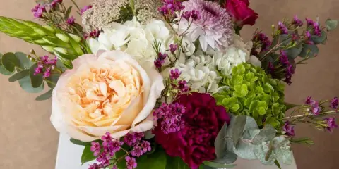 A mixed floral arrangement featuring peach roses, white hydrangeas, and purple accents in a square glass vase.