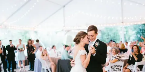 A bride and groom dancing at a wedding reception under a white tent with string lights.