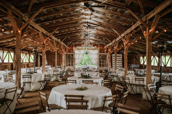 Interior of a rustic wooden barn venue with exposed beams, string lights, and round tables set for a wedding reception.