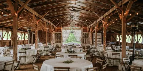 Interior of a rustic wooden barn venue with exposed beams, string lights, and round tables set for a wedding reception.