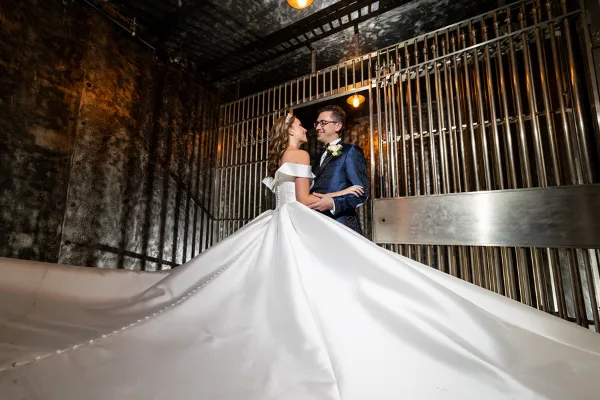 A bride and groom standing together in an industrial-style venue, with the bride's long white dress train spread out on the floor.