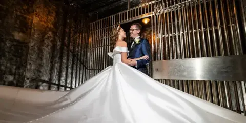 A bride and groom standing together in an industrial-style venue, with the bride's long white dress train spread out on the floor.