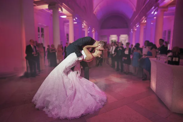 A bride and groom dancing at a wedding reception in a ballroom with pink lighting.