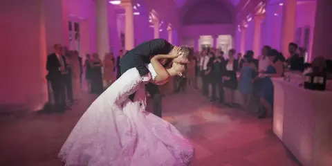 A bride and groom dancing at a wedding reception in a ballroom with pink lighting.