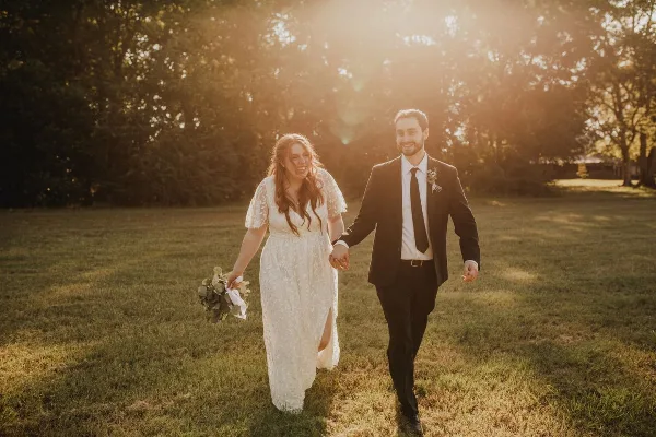 A bride and groom walking hand-in-hand through a sunlit grassy field.