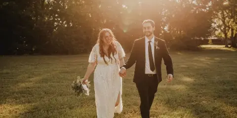 A bride and groom walking hand-in-hand through a sunlit grassy field.