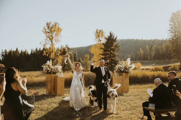 A bride and groom walking down the aisle at an outdoor wedding ceremony in a grassy field with a forest background.