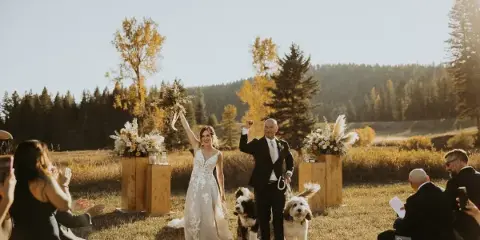 A bride and groom walking down the aisle at an outdoor wedding ceremony in a grassy field with a forest background.