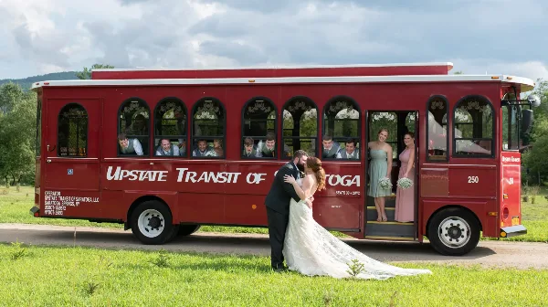 A red Upstate Transit of Saratoga trolley bus with a wedding party boarding and a couple kissing in front of it.