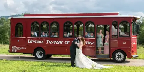 A red Upstate Transit of Saratoga trolley bus with a wedding party boarding and a couple kissing in front of it.