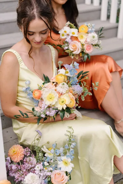 A bride and bridesmaid holding colorful wedding bouquets featuring roses, ranunculus, and delphinium.