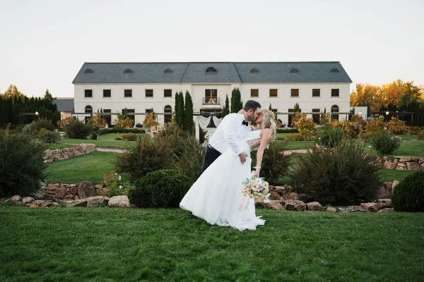 A bride and groom share a kiss on a lawn in front of a large white estate building.