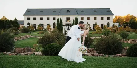 A bride and groom share a kiss on a lawn in front of a large white estate building.