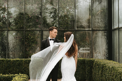 Bride and groom in wedding attire standing in front of a glass wall with greenery